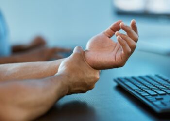 person in blue scrubs sitting at a desk and holding their left wrist with their right hand, indicating pain.