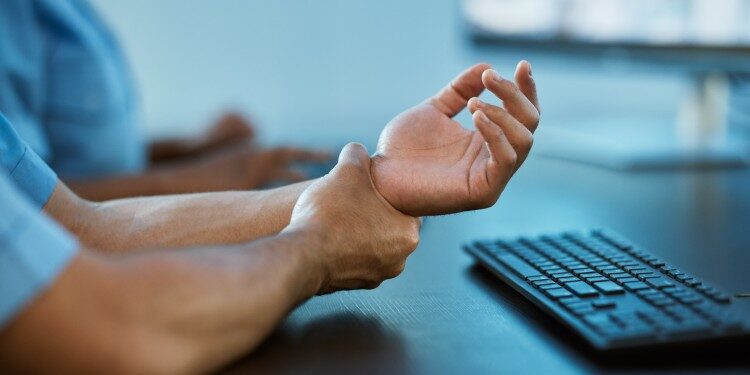 person in blue scrubs sitting at a desk and holding their left wrist with their right hand, indicating pain.