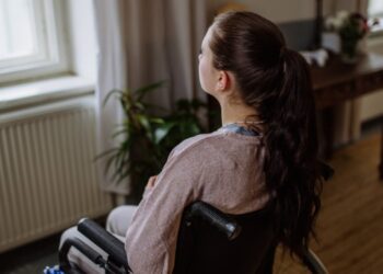 A young woman sitting in a wheelchair and looking out the window in her living room.
