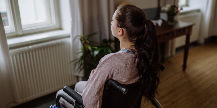 A young woman sitting in a wheelchair and looking out the window in her living room.