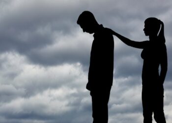 The silhouette of a couple on the beach under cloudy skies with one partner offering support to the other.