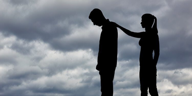 The silhouette of a couple on the beach under cloudy skies with one partner offering support to the other.