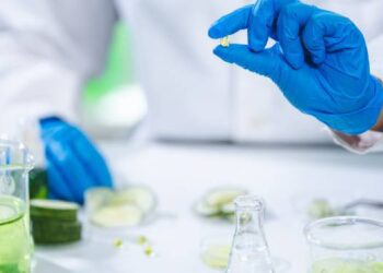A gloved hand holding a clear supplement pill over an experiment table with cucumber slices in beakers and Petri dishes.