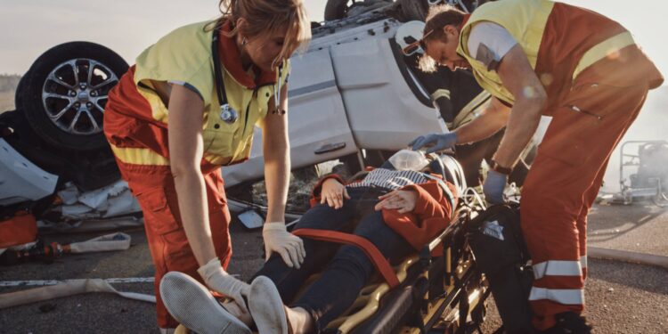 Two paramedics render emergency care to an injured person stabilized on a stretcher. A rolled-over vehicle lies behind them.