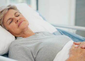 A woman lies in a hospital bed with closed eyes and a gentle smile. Her hands are on her abdomen, and her head is slightly raised.