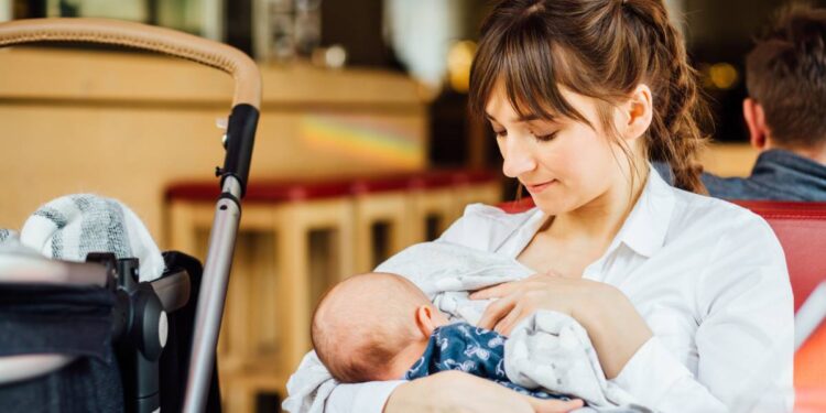 A young mother holding her infant at a dining table in public. She's confidently breastfeeding with the stroller next to her.