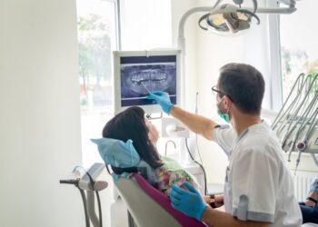 A licensed dentist pointing at an X-ray for a patient. The patient is sitting in the cleaning chair in a pink shirt.
