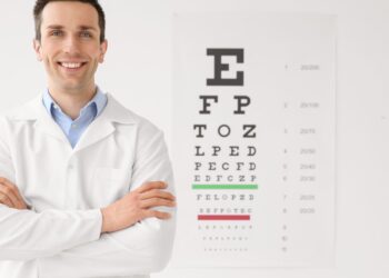 A smiling male eye doctor with his arms crossed, standing next to a vision chart that hangs on a white wall.