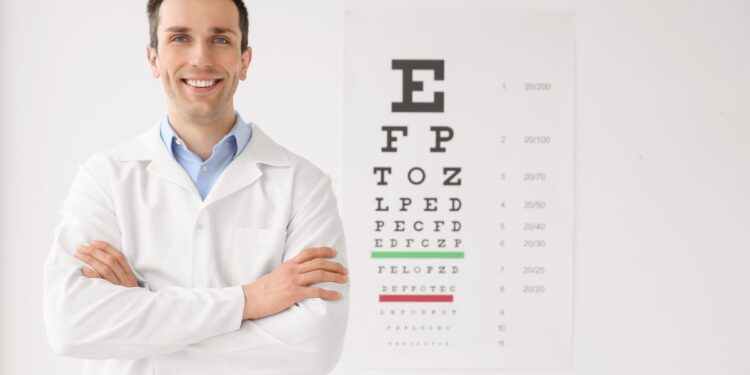 A smiling male eye doctor with his arms crossed, standing next to a vision chart that hangs on a white wall.
