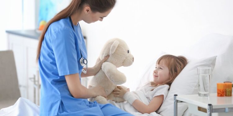 A doctor visiting a little girl in a hospital bed. The doctor is sitting at the edge of the bed holding a stuffed animal.