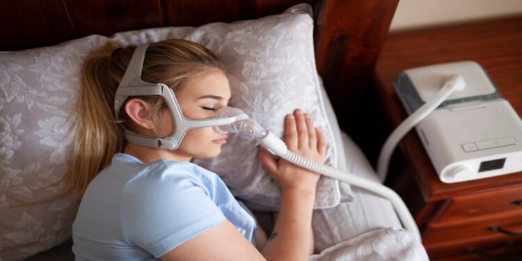 A woman wearing a blue shirt and a CPAP machine is sleeping on her side on a floral-patterned pillow.
