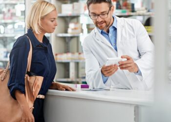 A pharmacist wearing a white coat is standing behind a counter and showing a female customer a box of medication.