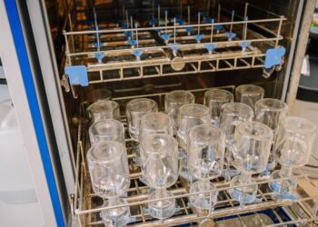 An open dishwasher surrounded by containers holding upside-down laboratory glassware as it hangs to dry.