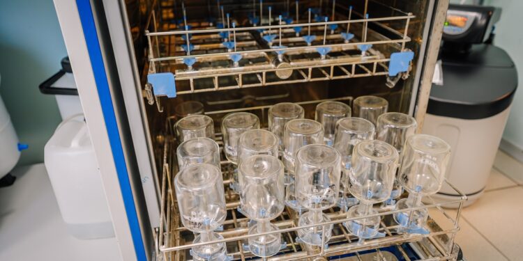 An open dishwasher surrounded by containers holding upside-down laboratory glassware as it hangs to dry.