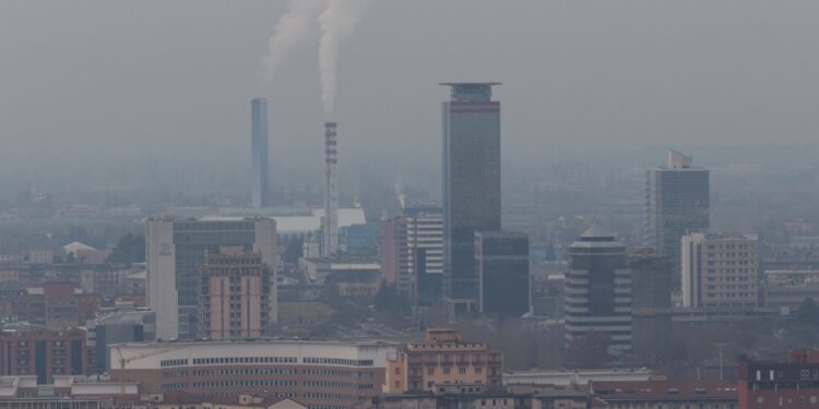 A cityscape with polluted air filling the sky. There's tall skinny smoke stacks with thick smoke releasing from the top.