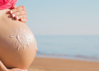 A pregnant woman in a bikini on a beach places her hands on her belly, which has a sun drawn on it with sunscreen.