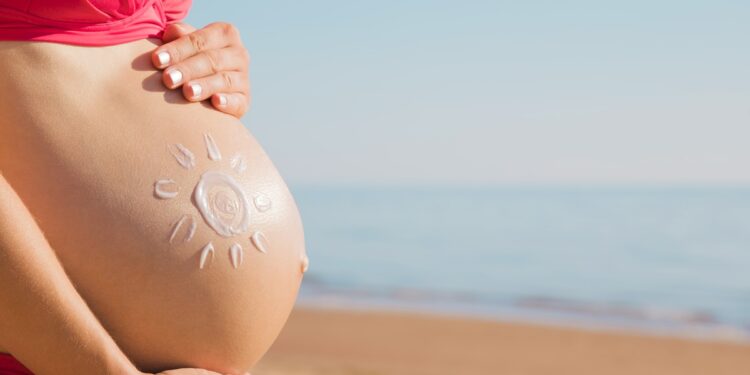 A pregnant woman in a bikini on a beach places her hands on her belly, which has a sun drawn on it with sunscreen.