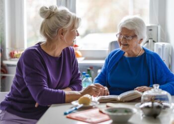 Two women, one older than the other, sit at a kitchen table smiling at each other. There is a book between them.