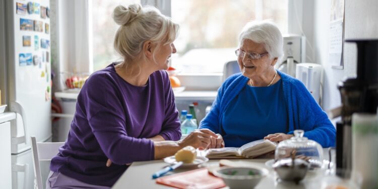 Two women, one older than the other, sit at a kitchen table smiling at each other. There is a book between them.
