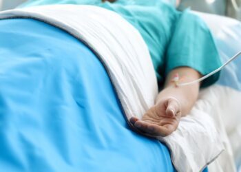 A medical patient laying in a hospital bed hooked up to an IV. They're in a teal green gown and a blue blanket.