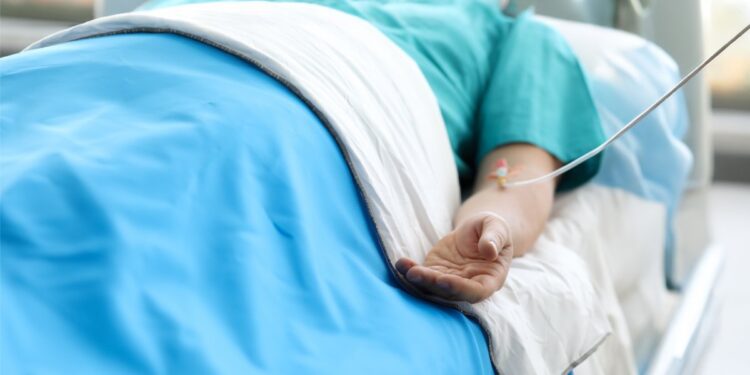 A medical patient laying in a hospital bed hooked up to an IV. They're in a teal green gown and a blue blanket.