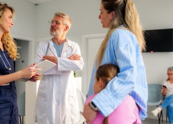 A woman hugs a young girl against her side while talking with two medical professionals in a waiting room.