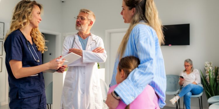 A woman hugs a young girl against her side while talking with two medical professionals in a waiting room.