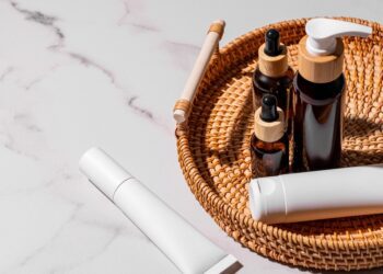 Amber glass bottles, white cream tubes, and skincare products are for daily use on a rattan tray with a marble background.