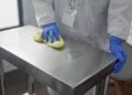 A lab professional wears blue latex gloves and a white lab coat to wipe down the stainless steel table surface.