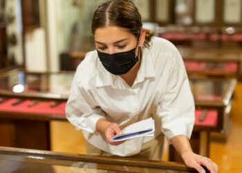 A young person learning over a museum display case while holding a booklet and wearing a black face mask.