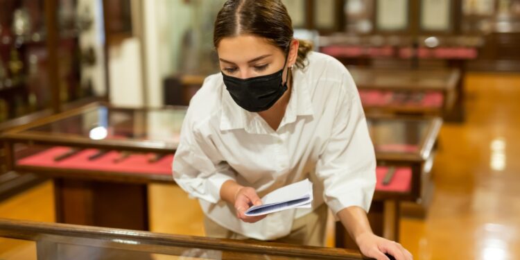 A young person learning over a museum display case while holding a booklet and wearing a black face mask.
