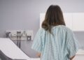 A woman wears a hospital gown and faces an exam table with a paper laid out. Other doctors' office supplies rest on the counter.