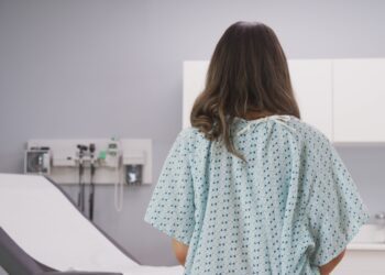 A woman wears a hospital gown and faces an exam table with a paper laid out. Other doctors' office supplies rest on the counter.