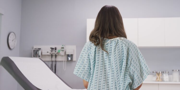 A woman wears a hospital gown and faces an exam table with a paper laid out. Other doctors' office supplies rest on the counter.