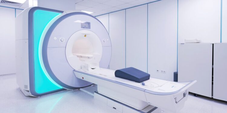 A high-tech MRI machine sits in a room at a hospital. A decorative panel on the side of the machine glows blue.