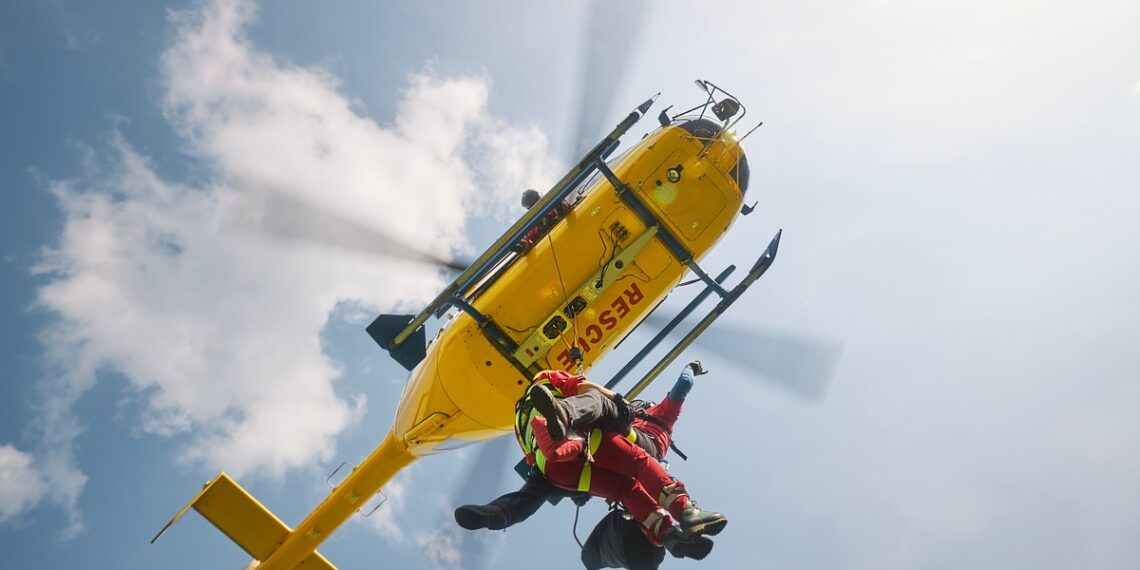 Two paramedics wearing red uniforms hang on a rope attached to a yellow helicopter that says 'Rescue' at the bottom.