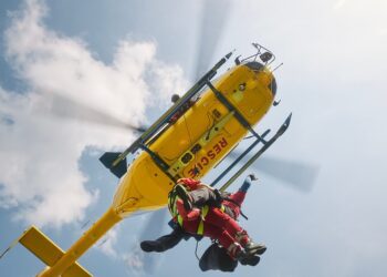 Two paramedics wearing red uniforms hang on a rope attached to a yellow helicopter that says 'Rescue' at the bottom.
