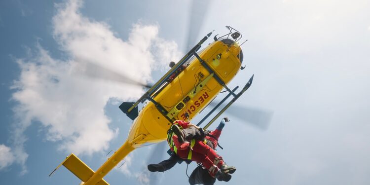 Two paramedics wearing red uniforms hang on a rope attached to a yellow helicopter that says 'Rescue' at the bottom.