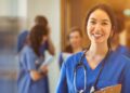 A woman wearing deep blue scrubs and a stethoscope smiles while holding a clipboard. Behind her is a group of people.