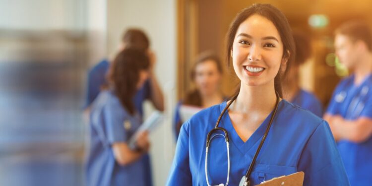 A woman wearing deep blue scrubs and a stethoscope smiles while holding a clipboard. Behind her is a group of people.