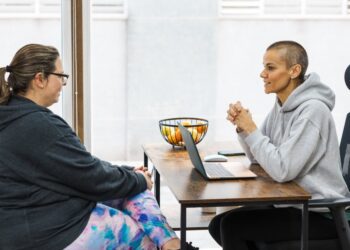 Two people wearing hoodies and leggings sit across from each other at a small desk near a large window.