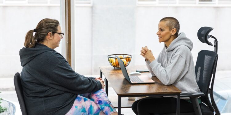 Two people wearing hoodies and leggings sit across from each other at a small desk near a large window.