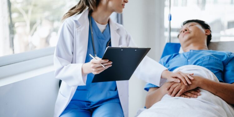 A female nurse wearing blue scrubs and a white coat sits next to a patient lying in bed, with her hand on his clasped hands.