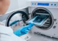 A healthcare professional wears a blue latex glove as they place a sterilizing tray into an autoclave device.