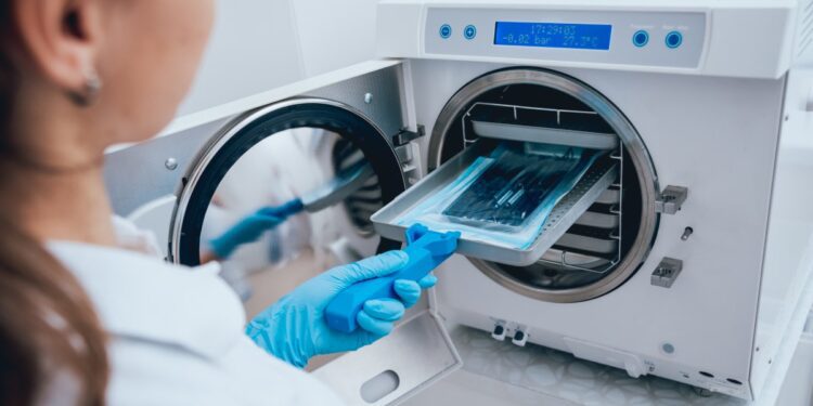 A healthcare professional wears a blue latex glove as they place a sterilizing tray into an autoclave device.