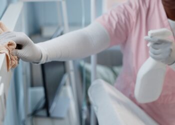 A hospital employee cleans surfaces in a patient's recovery room with a bottle of cleaning solution and a rag.