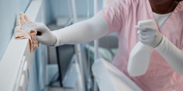 A hospital employee cleans surfaces in a patient's recovery room with a bottle of cleaning solution and a rag.