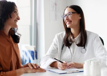 Woman sitting across from a female doctor behind a desk writing on a clipboard next to a computer inside a clinic.