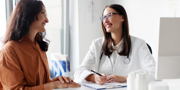 Woman sitting across from a female doctor behind a desk writing on a clipboard next to a computer inside a clinic.