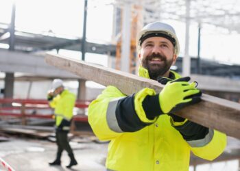 A smiling man wearing a white hard hat, yellow high visibility coat, and yellow high visibility gloves is carrying a long piece of lumber on his shoulder at a construction site.